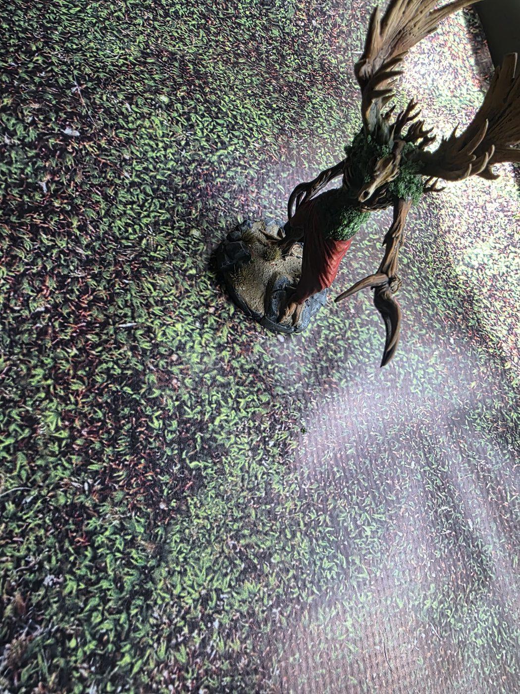 The tree-creature mini on the moss mat from above, a white patch of window glare across the right side