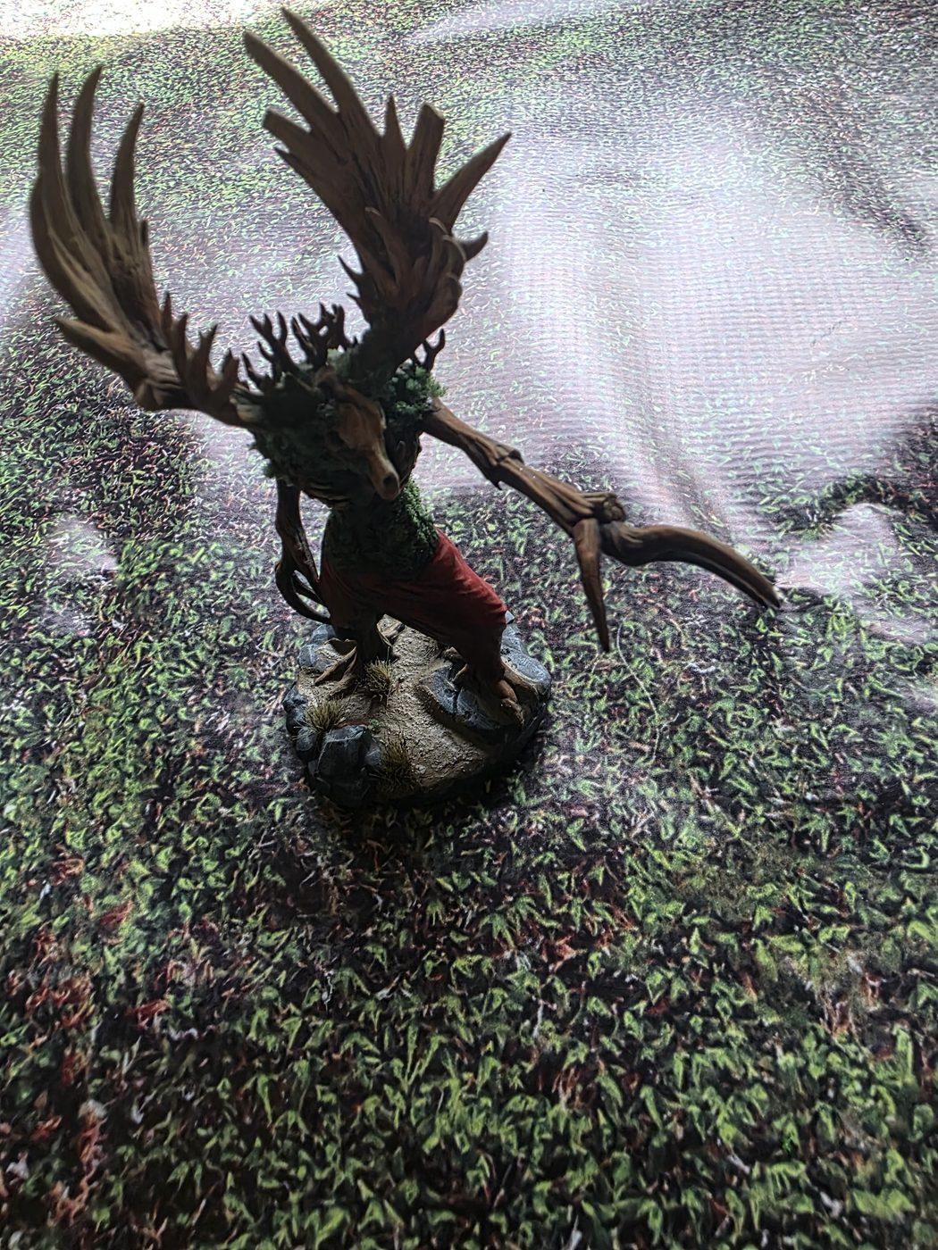 A painted tree-creature miniature standing on the moss print mat, with glare washing out the print behind it